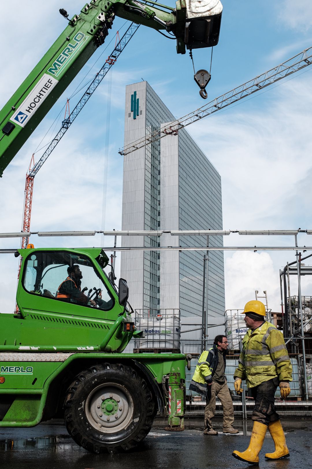 Grünes Kranfahrzeug und Bauarbeiter in gelb vor Hochhaus